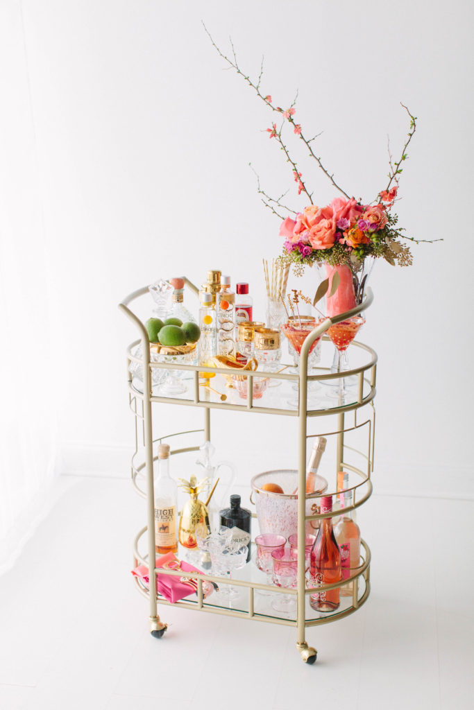 punchy styled bar cart by The Prissy Plate Company, with coral vase, orange cosmopolitans, a gold bowl of limes, and rose and champagne bottles